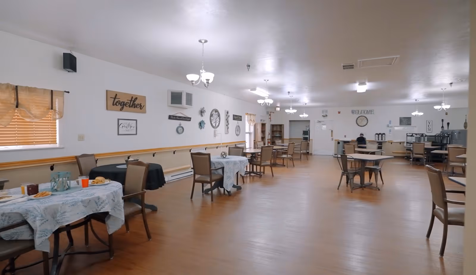 A spacious dining room with several tables covered with tablecloths and chairs arranged around them. The room has wooden flooring, white walls decorated with various wall art and signs, and multiple ceiling light fixtures. There is a window with blinds on the left side and a counter area with coffee dispensers and other items at the far end of the room.