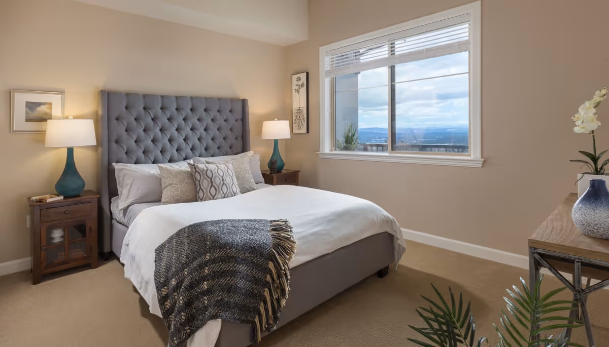 A neatly made bed with a tufted gray headboard in a bedroom. The bed has white sheets, several decorative pillows, and a dark gray throw blanket. There are two wooden nightstands on either side of the bed, each with a teal lamp. A window on the right side shows a scenic view of mountains and sky. The room has beige walls and carpet, with framed artwork on the walls and a wooden dresser with a blue and white vase and a plant.