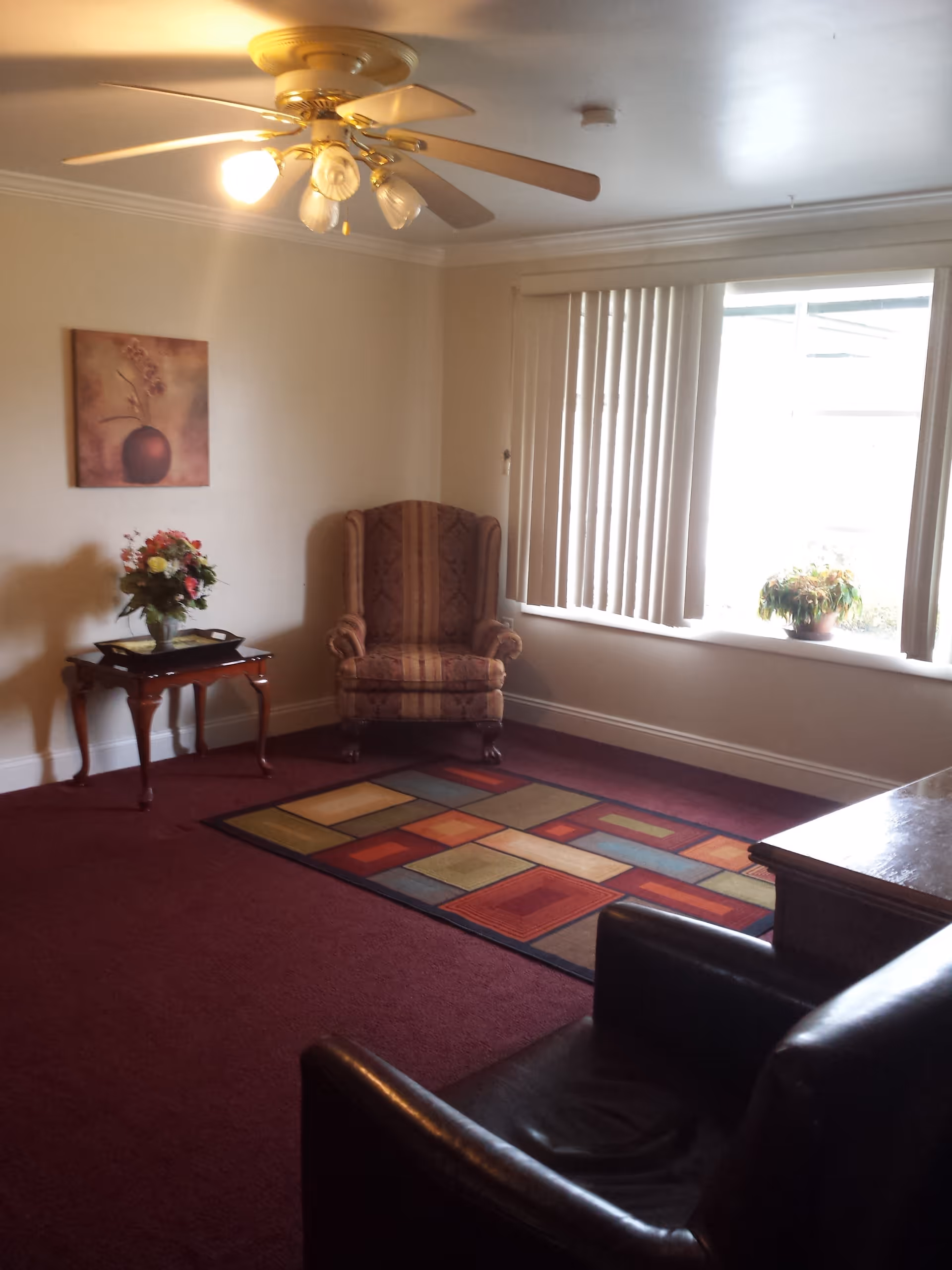 A cozy living room with a ceiling fan and light fixture, a patterned armchair, a small wooden side table with a floral arrangement, a colorful geometric area rug on a maroon carpet, a large window with vertical blinds, and a dark leather chair partially visible in the foreground.