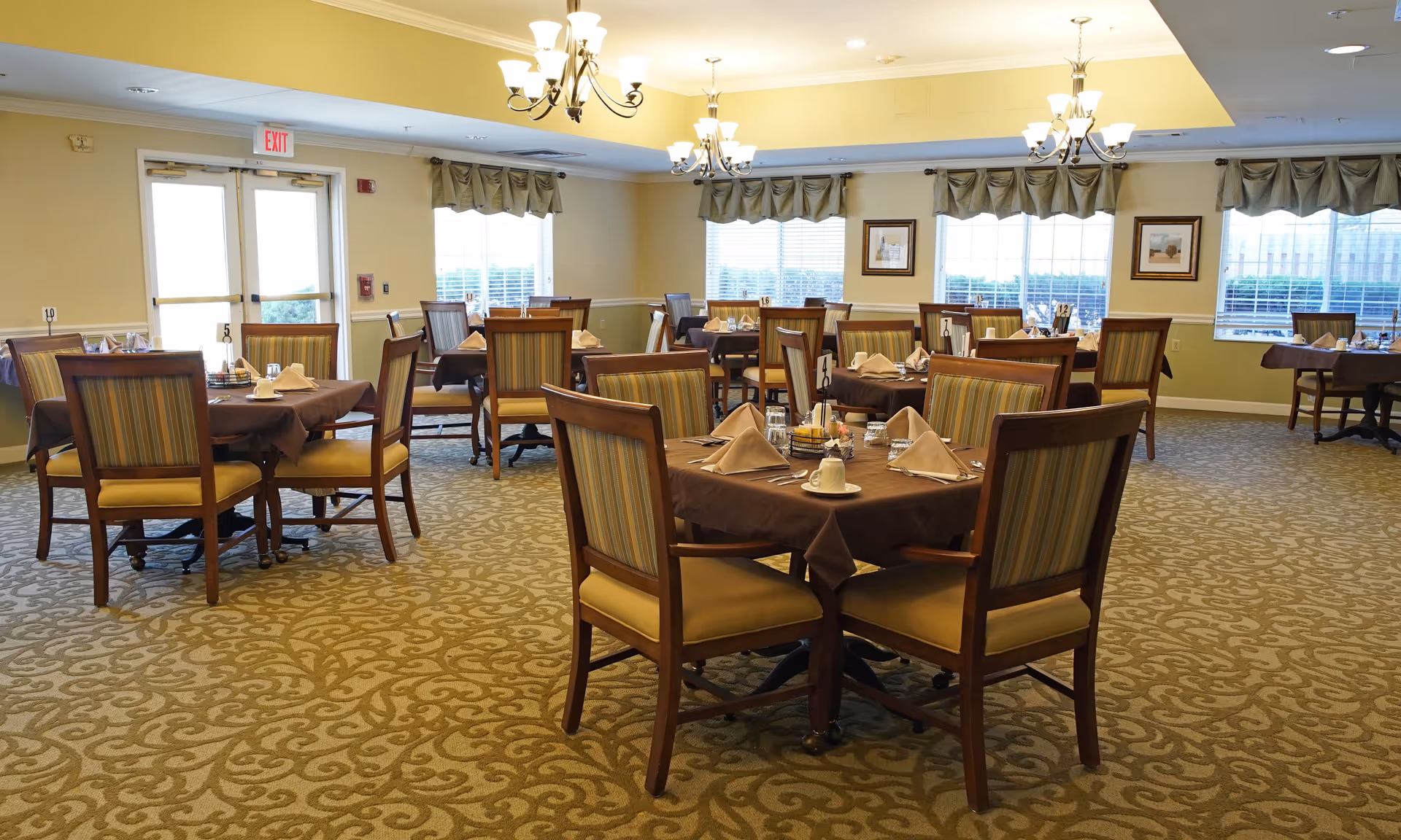 A spacious dining room with multiple tables covered in brown tablecloths, each set with napkins, cups, and utensils. The room has patterned carpet, large windows with valances, chandeliers hanging from the ceiling, and framed pictures on the walls.