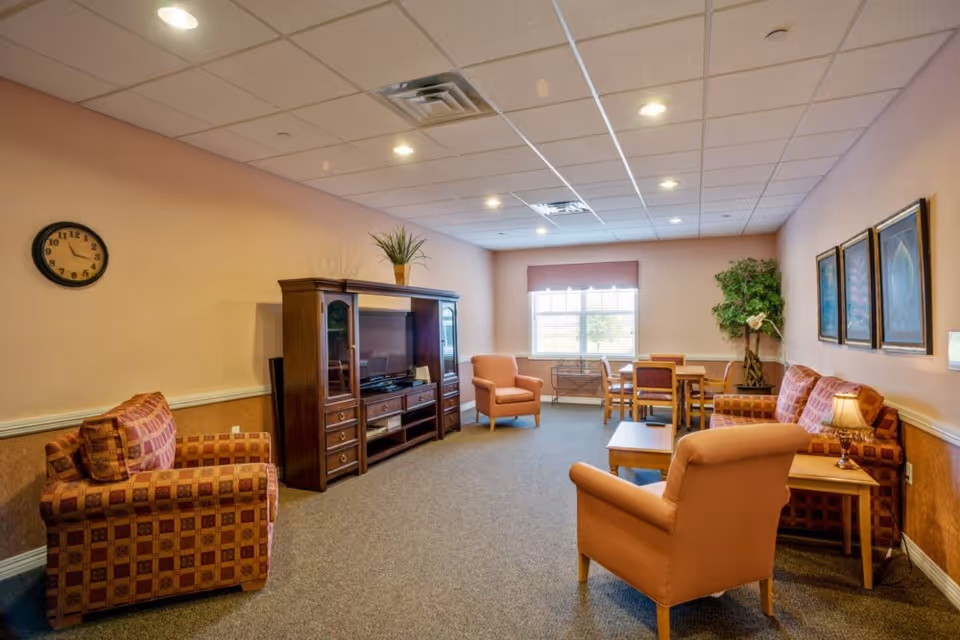 A bright senior living common room with upholstered chairs and sofas, a TV cabinet, tables, and a window letting in daylight.
