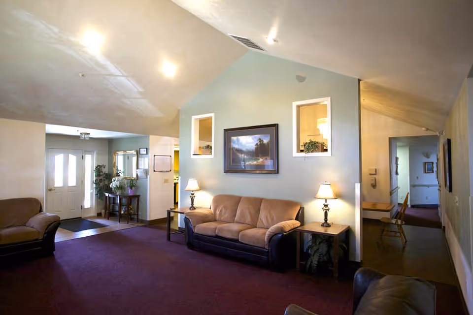 A cozy living room area with a beige couch and two side tables with lamps on either side. The walls are painted light green and decorated with a framed landscape painting and two small windows with plants. There is a purple carpet covering the floor, and the room has a high, slanted ceiling with recessed lighting. In the background, there is a hallway and an entrance door with glass panels, along with a small table holding plants and a mirror above it.