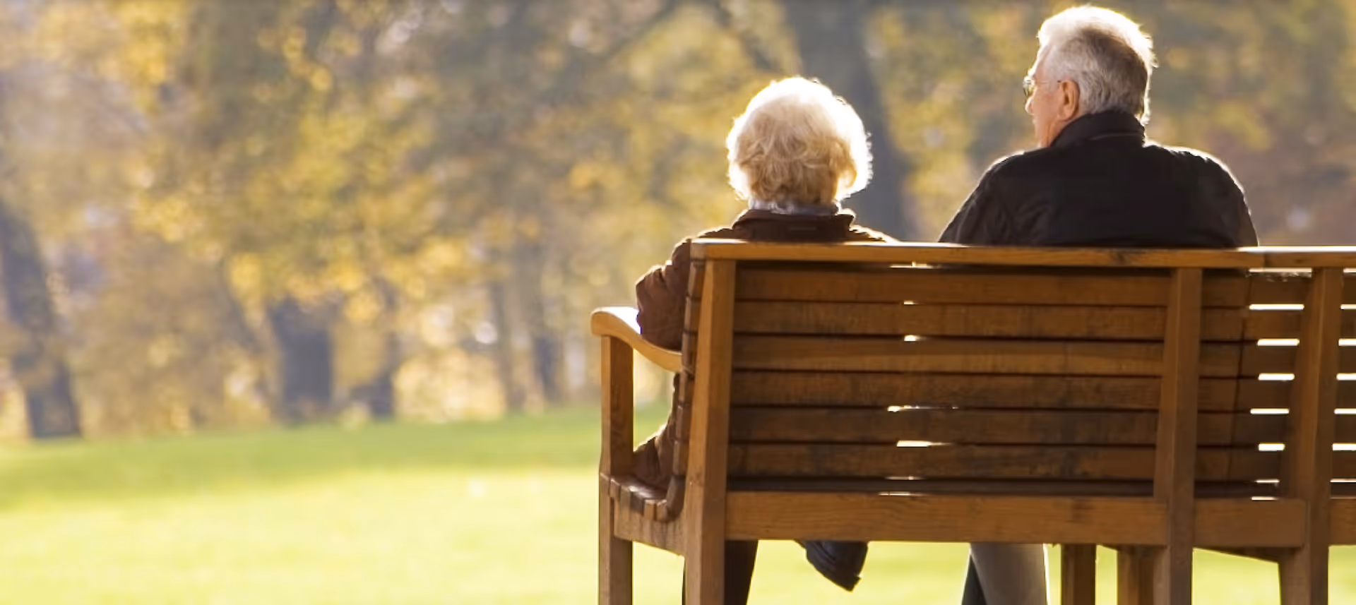 Two elderly individuals sitting on a wooden bench in a park, facing away from the camera with trees and greenery in the background.