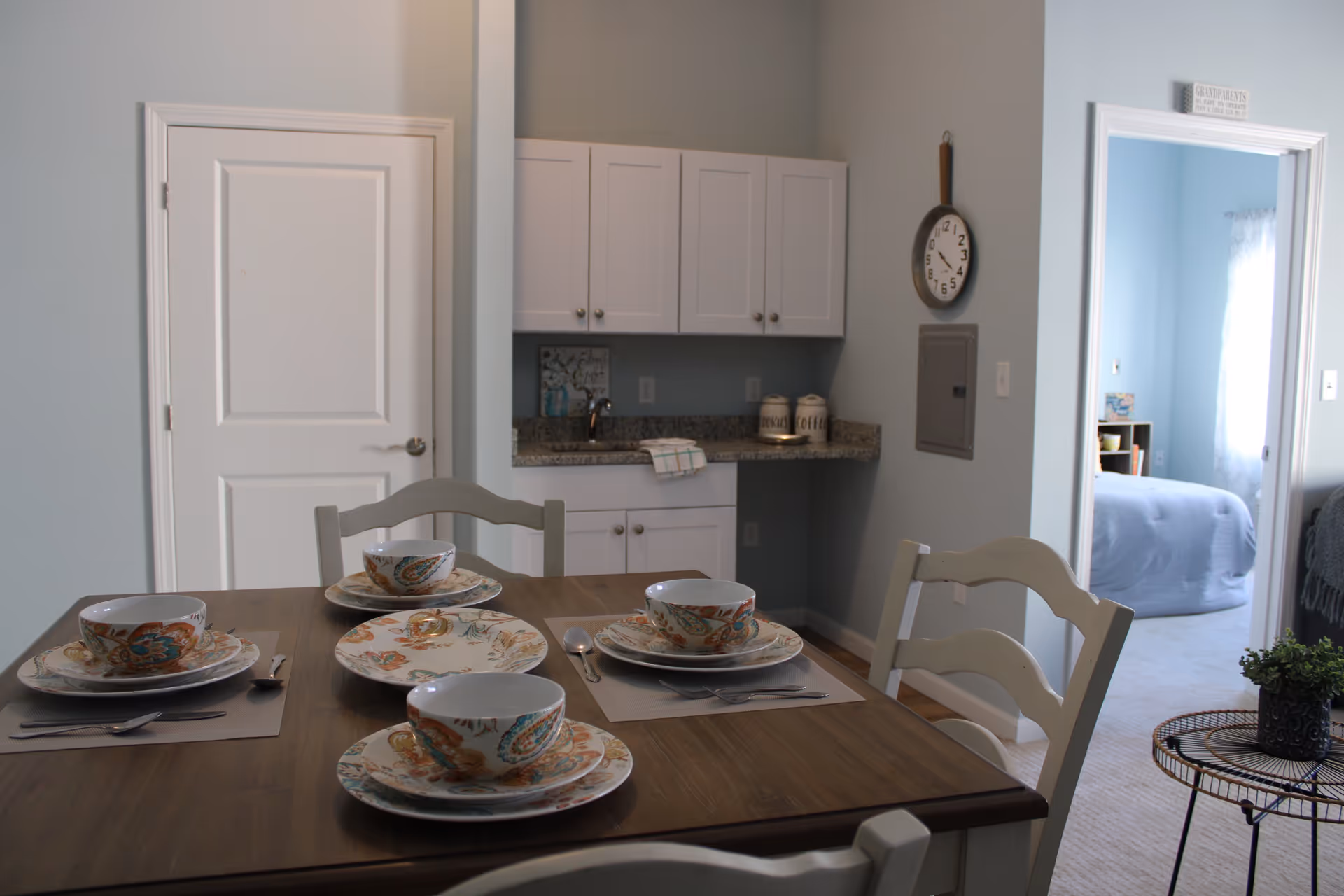 A dining area with a wooden table set for four with floral patterned dishes and bowls. In the background, there is a small kitchenette with white cabinets and a granite countertop. A clock hangs on the wall next to an electrical panel. To the right, an open doorway leads to a bedroom with a bed covered in a blue blanket and a window with sheer curtains.