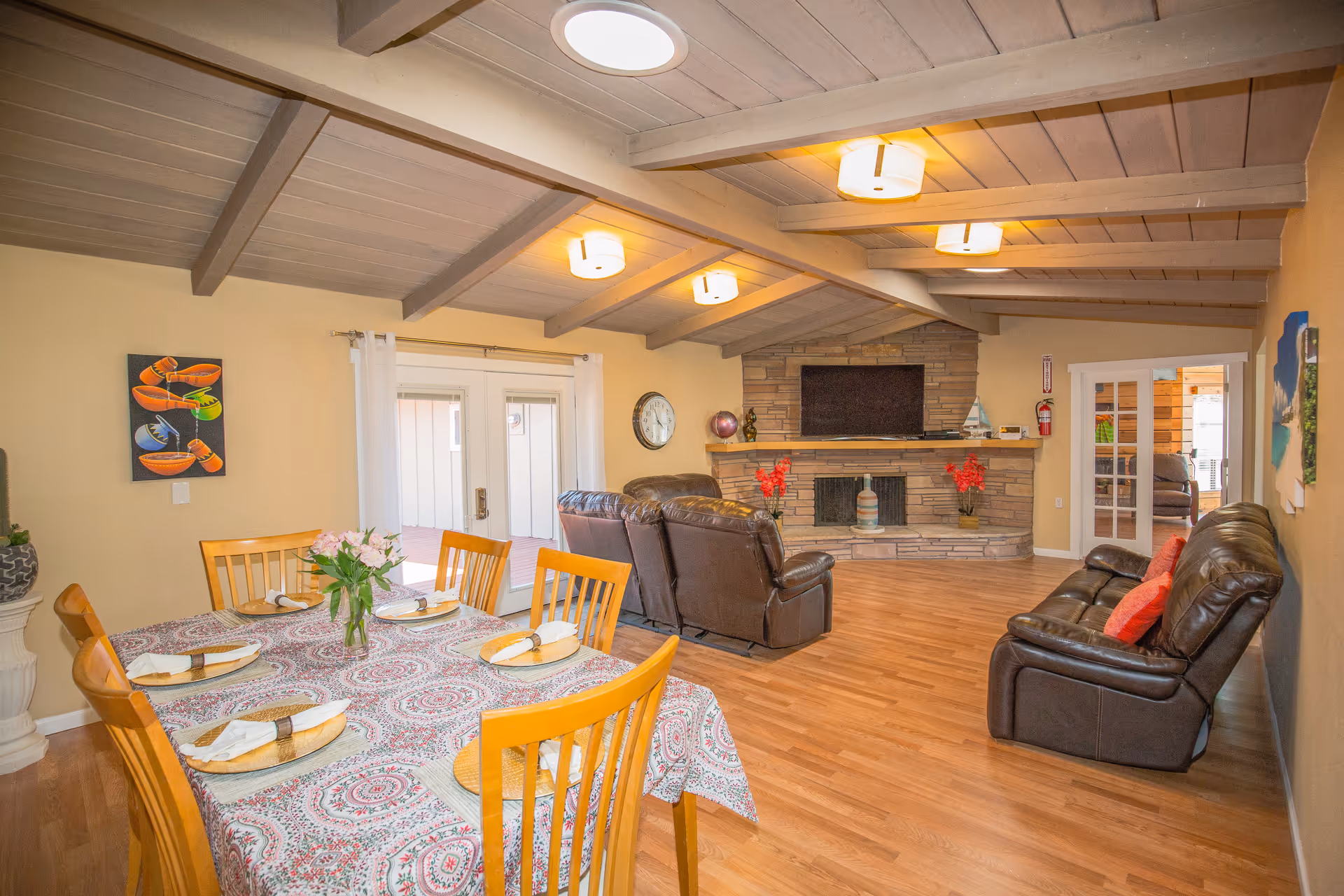 A spacious living and dining area with wooden floors and a vaulted ceiling with exposed beams. The dining table is set with six wooden chairs, a patterned tablecloth, and a vase of flowers. The living area features two brown leather sofas and a matching recliner facing a stone fireplace with a mounted flat-screen TV. There are decorative items on the fireplace mantel and wall art on the walls. French doors lead to another room.