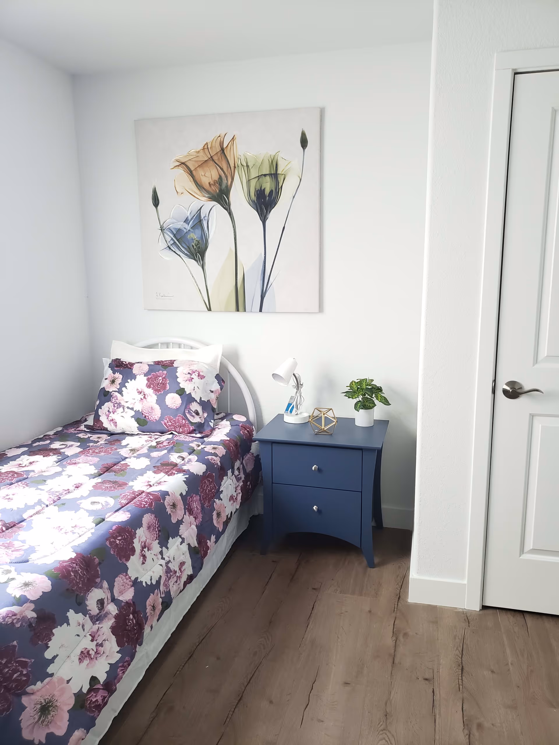 A small bedroom with a single bed covered in a floral purple and white bedspread and matching pillow. Next to the bed is a blue nightstand with two drawers, holding a small white lamp, a decorative geometric object, and a small potted plant. Above the bed hangs a painting of three large flowers in soft colors. The room has light-colored walls and wooden flooring.