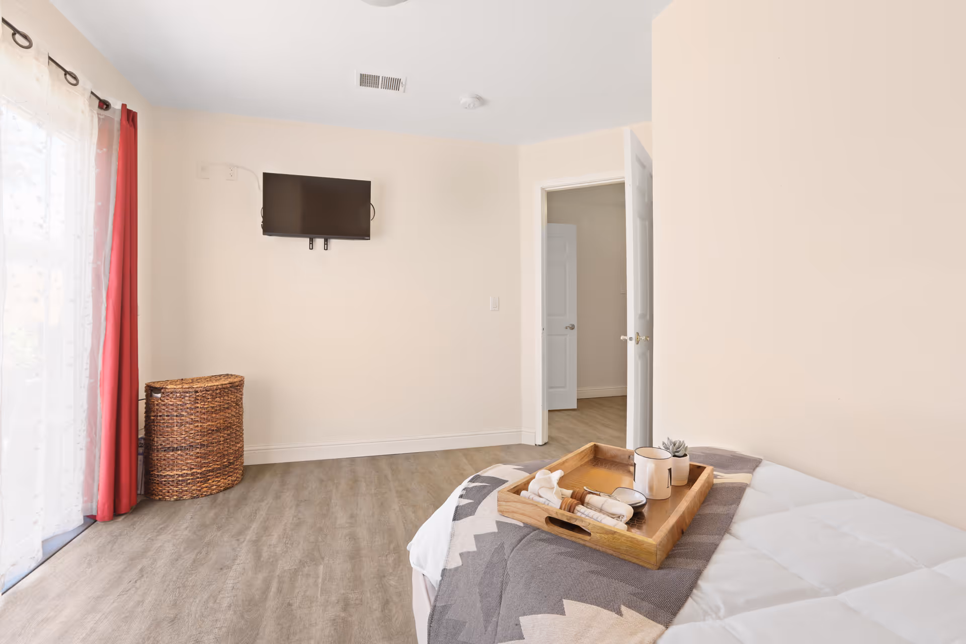 A bedroom with a bed partially visible in the foreground, topped with a wooden tray holding a cup, a small plant, and some rolled napkins. The room has light-colored walls, a wall-mounted flat-screen TV, a wicker basket in the corner, and a door leading to another room. There is a window with sheer white curtains and red drapes on the left side.