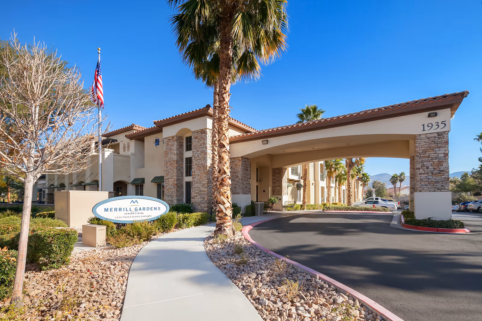 Exterior view of Merrill Gardens at Green Valley Ranch senior living facility entrance with a covered driveway, palm trees, landscaping, and a clear blue sky.