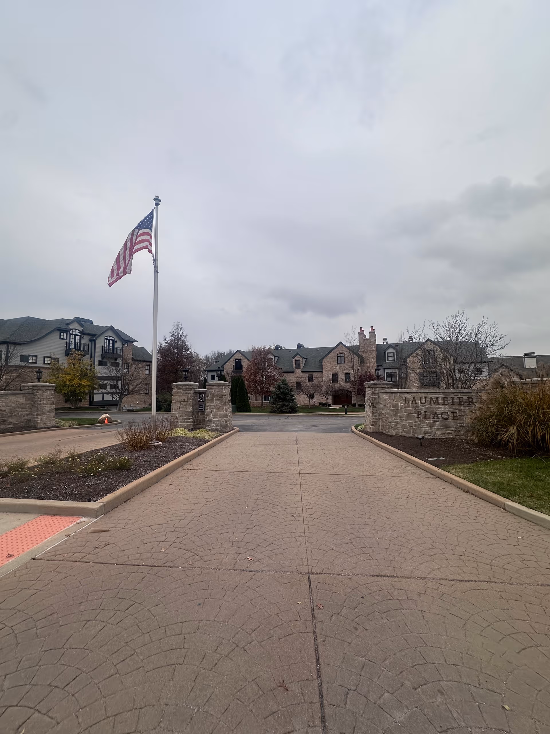 Entrance driveway to a residential facility with stone pillars and a flagpole flying the American flag. The building in the background has a stone facade with multiple windows and chimneys. A stone sign on the right reads 'LAUMEIER PLACE'. The sky is overcast.