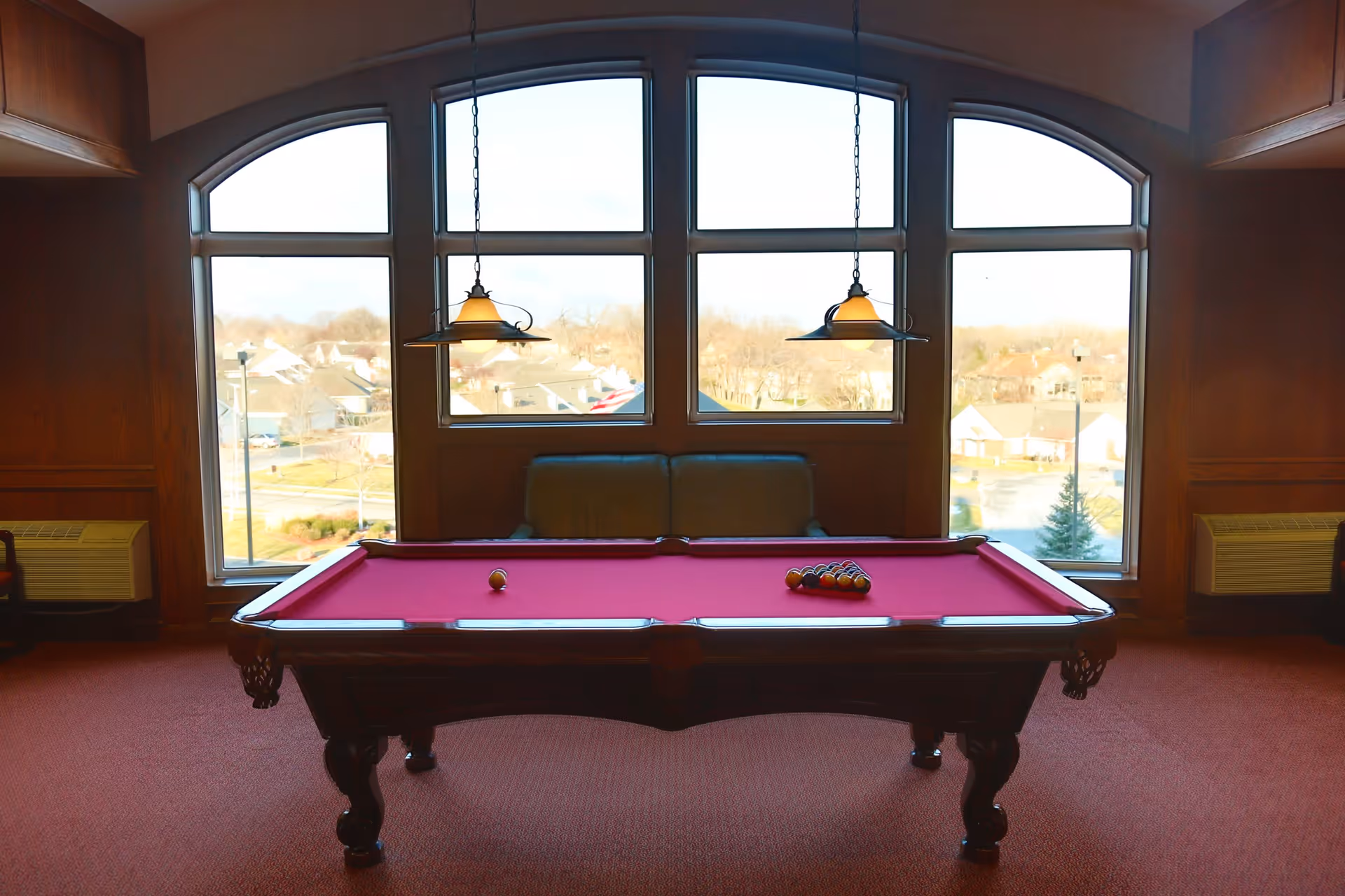 A pool table with a red felt surface is positioned in front of large arched windows showing a suburban neighborhood outside. Two pendant lights hang above the pool table, and a green couch is placed against the wall beneath the windows. The room has wood-paneled walls and red carpeting.