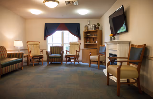 A cozy living room area in a senior living facility with several chairs arranged around the room. There is a window with a dark valance, a wooden bookshelf with decorative items, a wall-mounted TV above a white electric fireplace, and soft lighting from ceiling fixtures and a floor lamp.