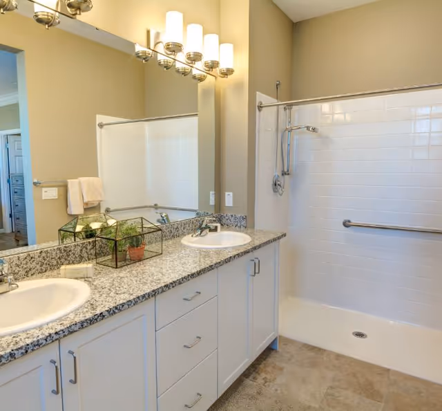 Bright bathroom with a double-sink granite vanity, large mirror, and a walk-in tiled shower with a grab bar.