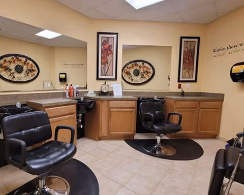 Interior view of a senior living facility's hair salon area featuring two black salon chairs on black mats, wooden cabinets with countertops, two decorative framed wall art pieces, a wall-mounted soap dispenser, and a large mirror reflecting the room.