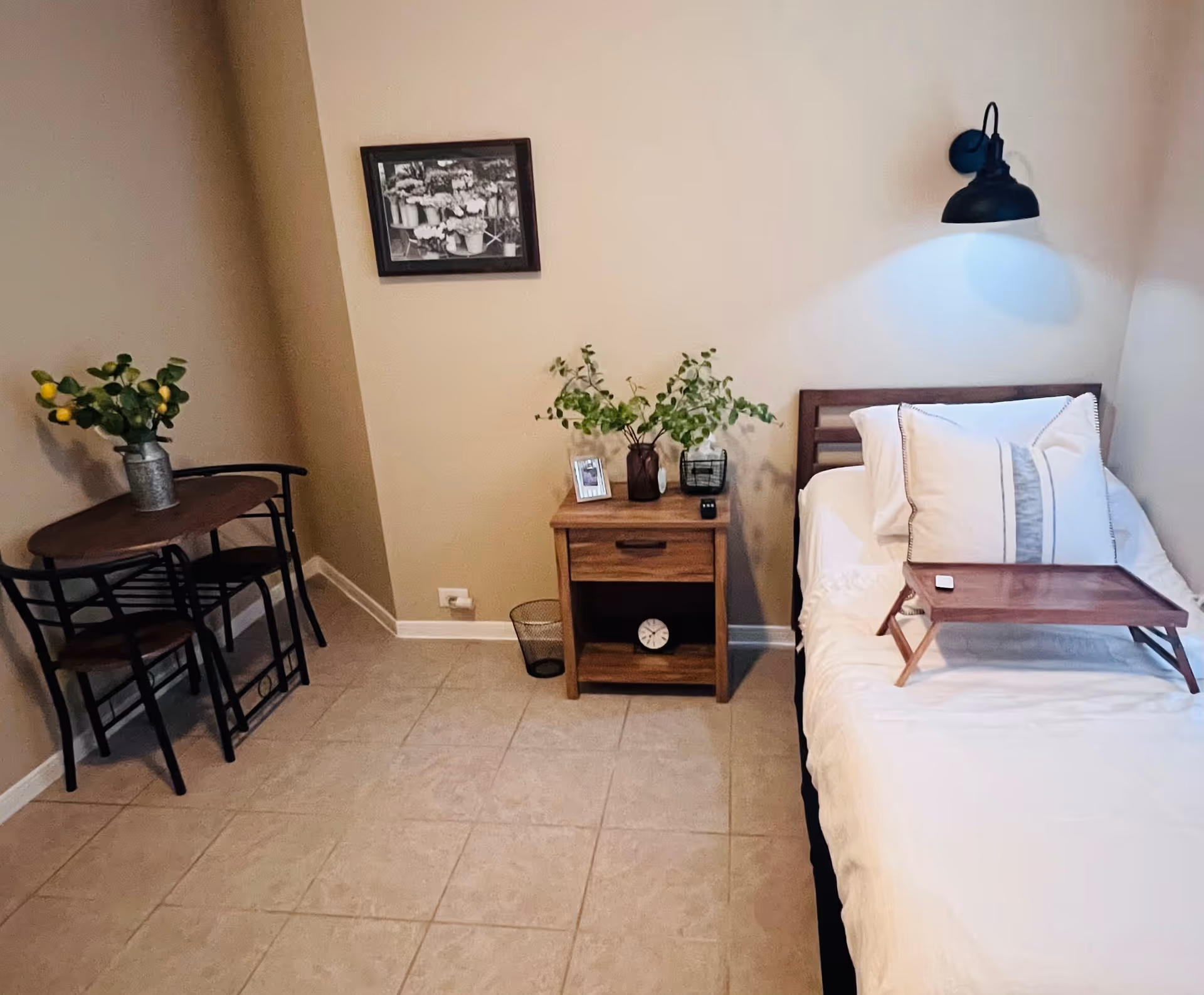Small furnished bedroom with a single bed, wooden nightstand, wall lamp, and a small table with chairs.