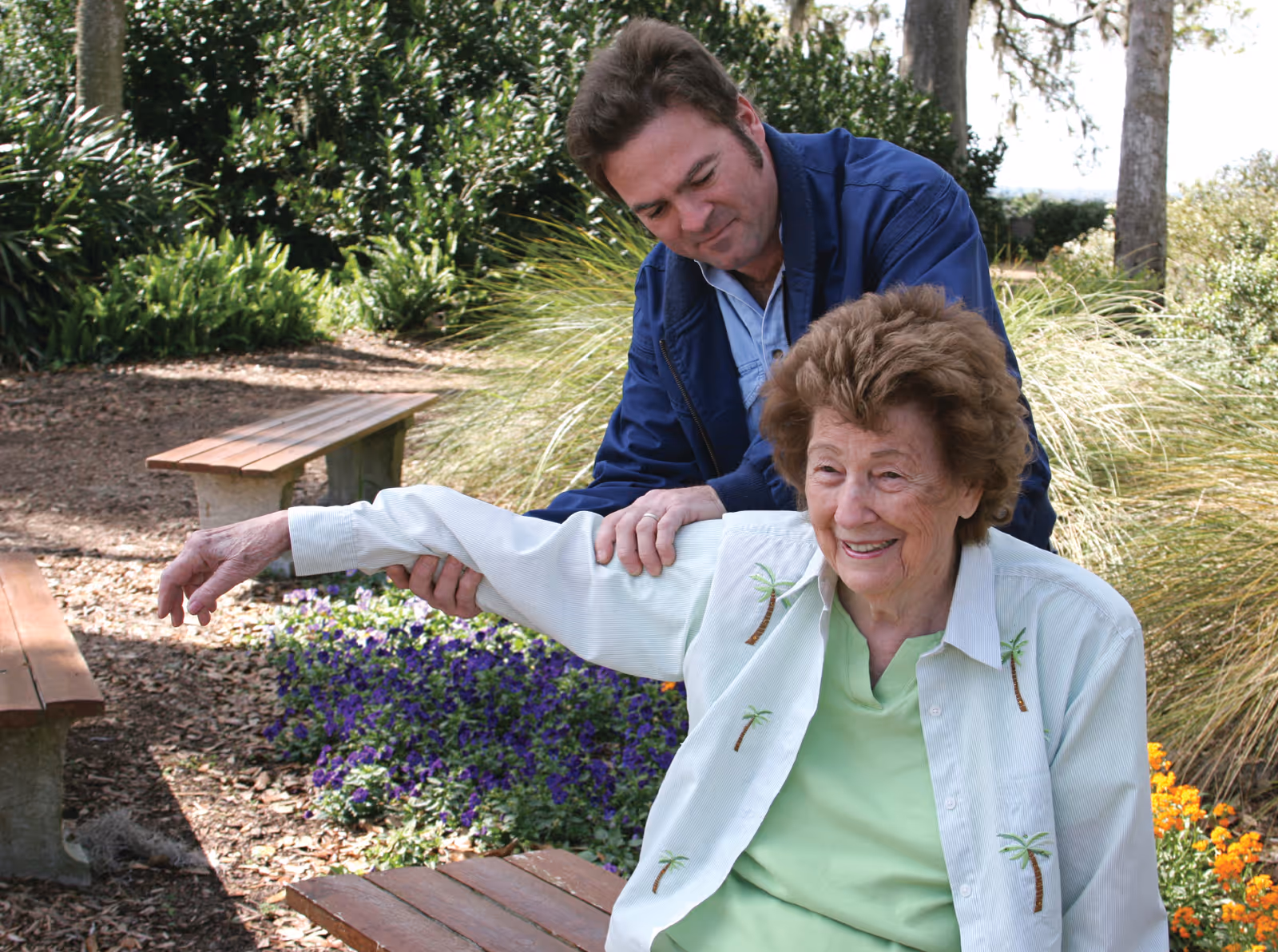An elderly woman sitting on a wooden bench outdoors, smiling as a man in a blue jacket assists her by gently stretching her arm. They are surrounded by greenery, flowers, and trees in a garden setting.