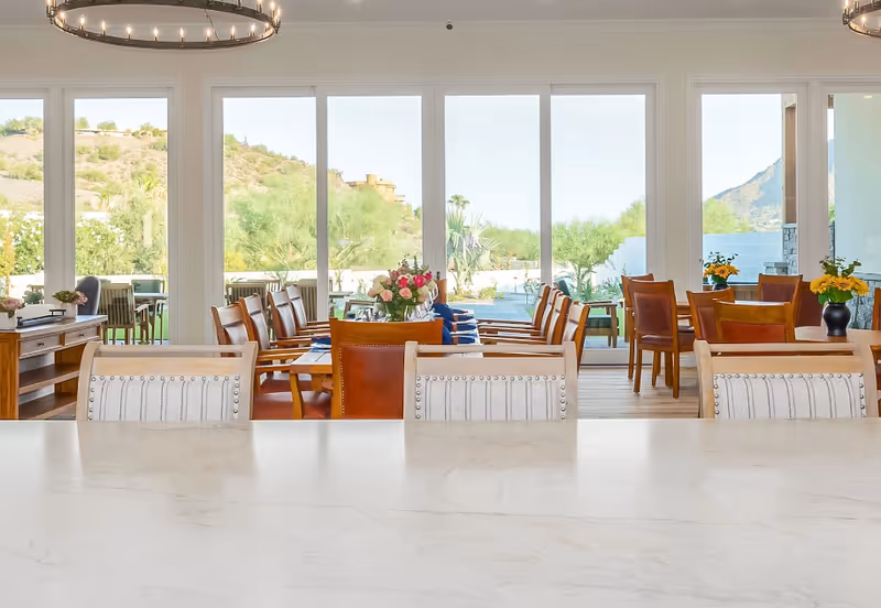 Bright dining room with multiple wooden tables and chairs, decorated with flower arrangements. Large floor-to-ceiling windows provide a view of an outdoor garden area with desert landscaping and mountains in the background.