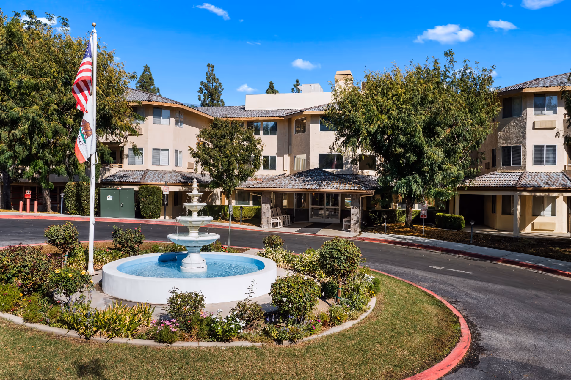 Exterior view of Columbus Estates senior living community featuring a circular driveway with a central water fountain surrounded by greenery and flowers, an American flag and California state flag on a flagpole, and a three-story beige building with multiple windows and a covered entrance under a clear blue sky.