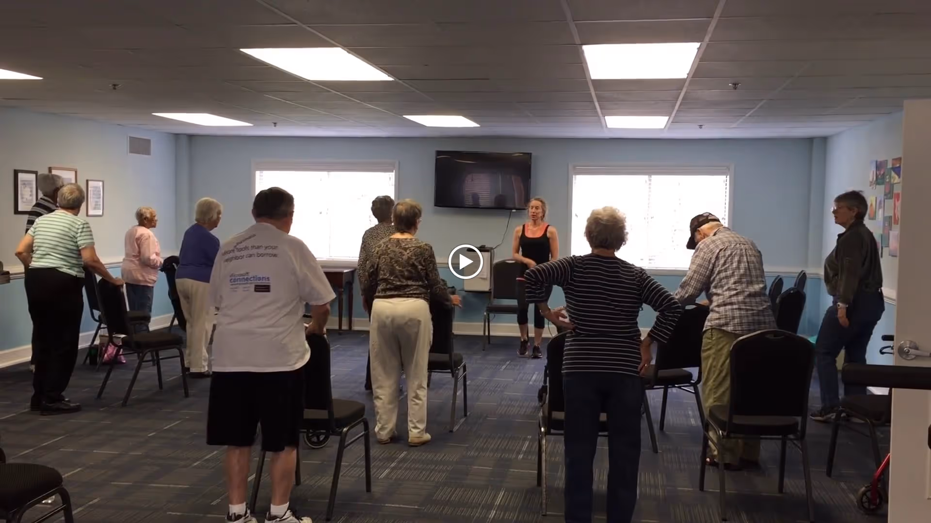 A group of elderly people participating in a seated exercise class in a community room with chairs arranged in rows. An instructor stands at the front of the room leading the session. The room has light blue walls, two windows, and a wall-mounted TV.