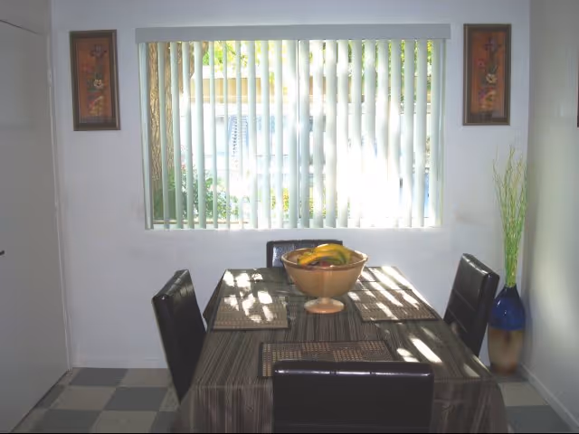 A small dining area with a table covered by a striped tablecloth and four black chairs. On the table is a wooden bowl filled with bananas and other fruit. Behind the table is a window with vertical blinds partially open, letting in sunlight. On either side of the window are framed floral pictures, and to the right is a tall blue vase with green decorative grass.