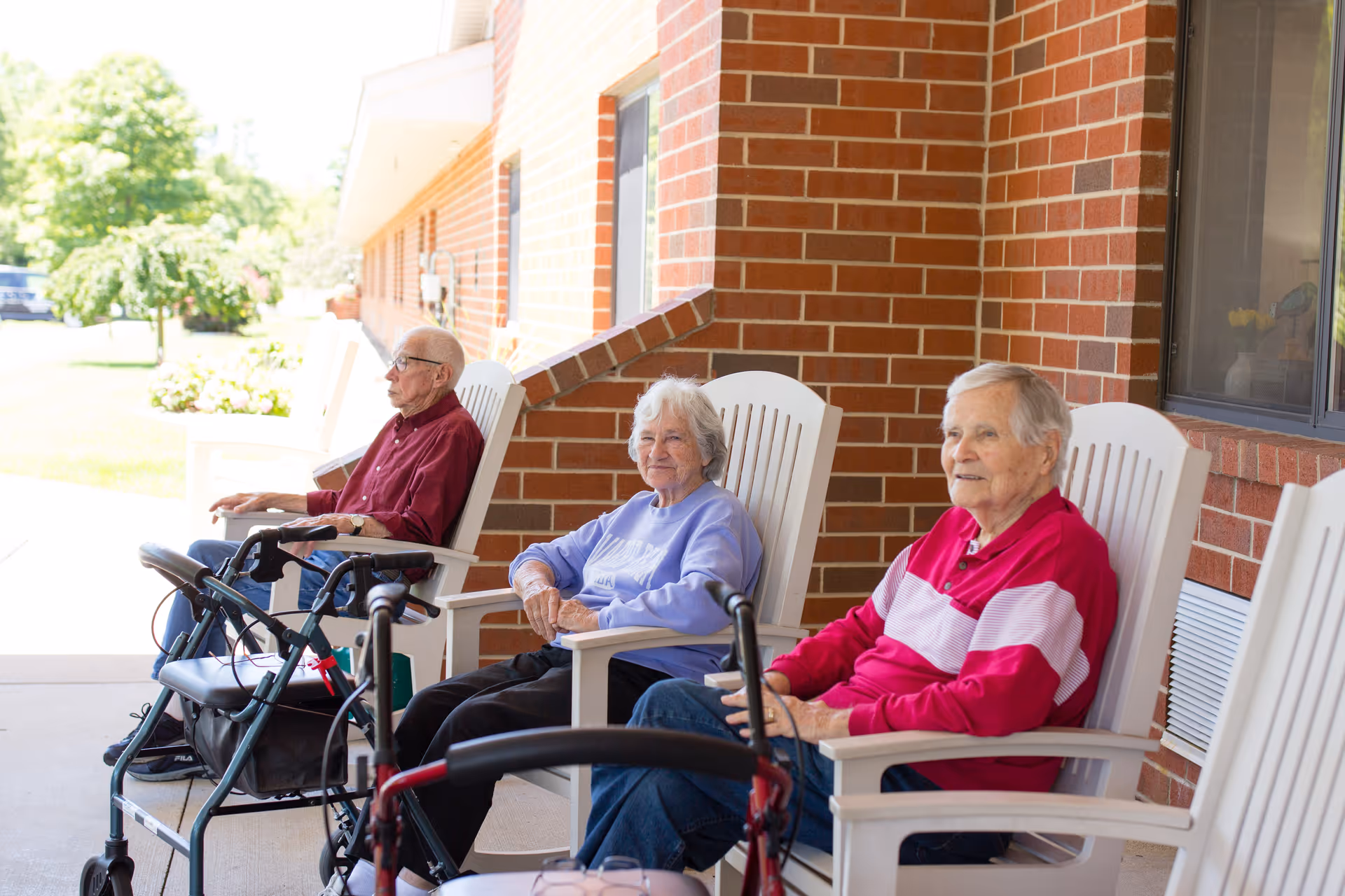 Three elderly individuals sitting on white chairs on a covered patio outside a brick building. Two walkers are positioned in front of them. The area is shaded and there is greenery visible in the background.