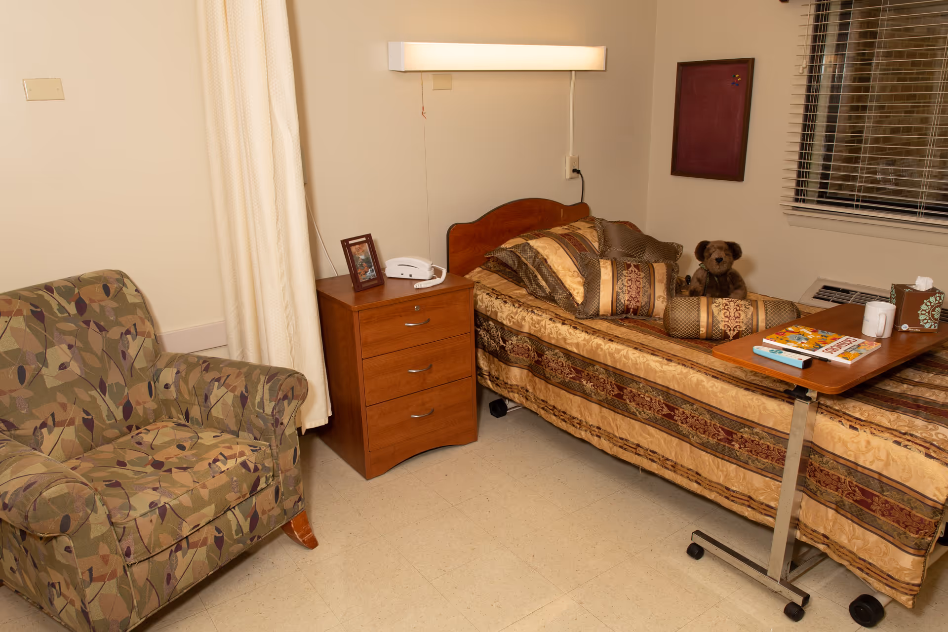 A small bedroom in a senior living facility featuring a single bed with patterned bedding, a teddy bear, and several pillows. Next to the bed is a wooden nightstand with a telephone and a framed photo. A patterned armchair is positioned to the left, and a rolling overbed table with books, a mug, and a tissue box is placed over the bed. A window with blinds and a wall-mounted light fixture are visible.