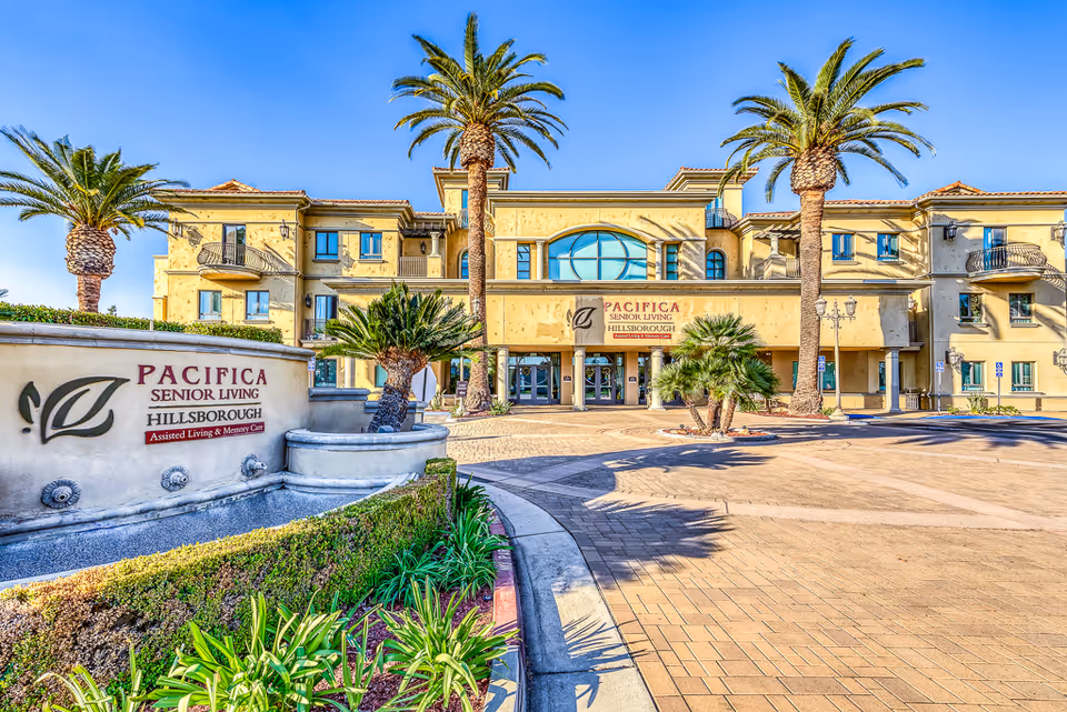 Exterior view of a senior living facility named Pacifica Senior Living Hillsborough with a large building featuring balconies, palm trees, and a clear blue sky. There is a sign in front with the facility's name and description as Assisted Living & Memory Care.