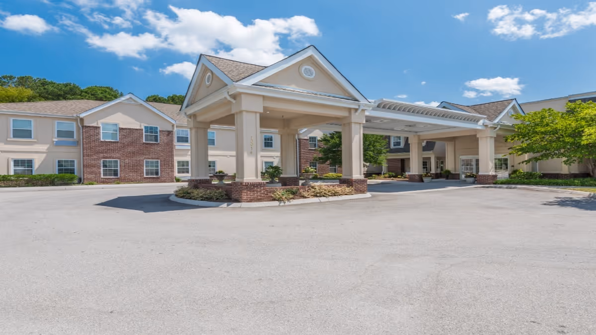 Exterior view of Park Place of West Knoxville senior living facility showing a covered entrance with columns, a driveway, and a two-story building with brick and beige siding under a blue sky with some clouds.
