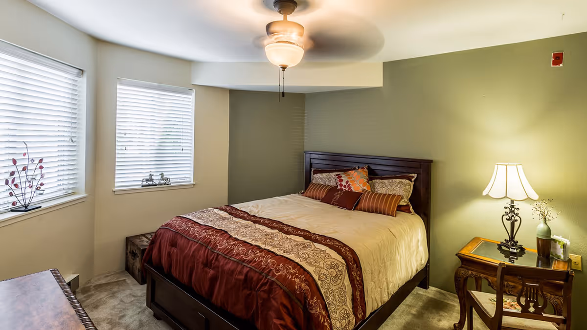 A cozy bedroom with a large bed featuring a decorative comforter and multiple pillows. The room has two windows with blinds, a ceiling fan with a light, a wooden nightstand with a lamp and a small vase, and a wooden chair. The walls are painted in light beige and green tones.