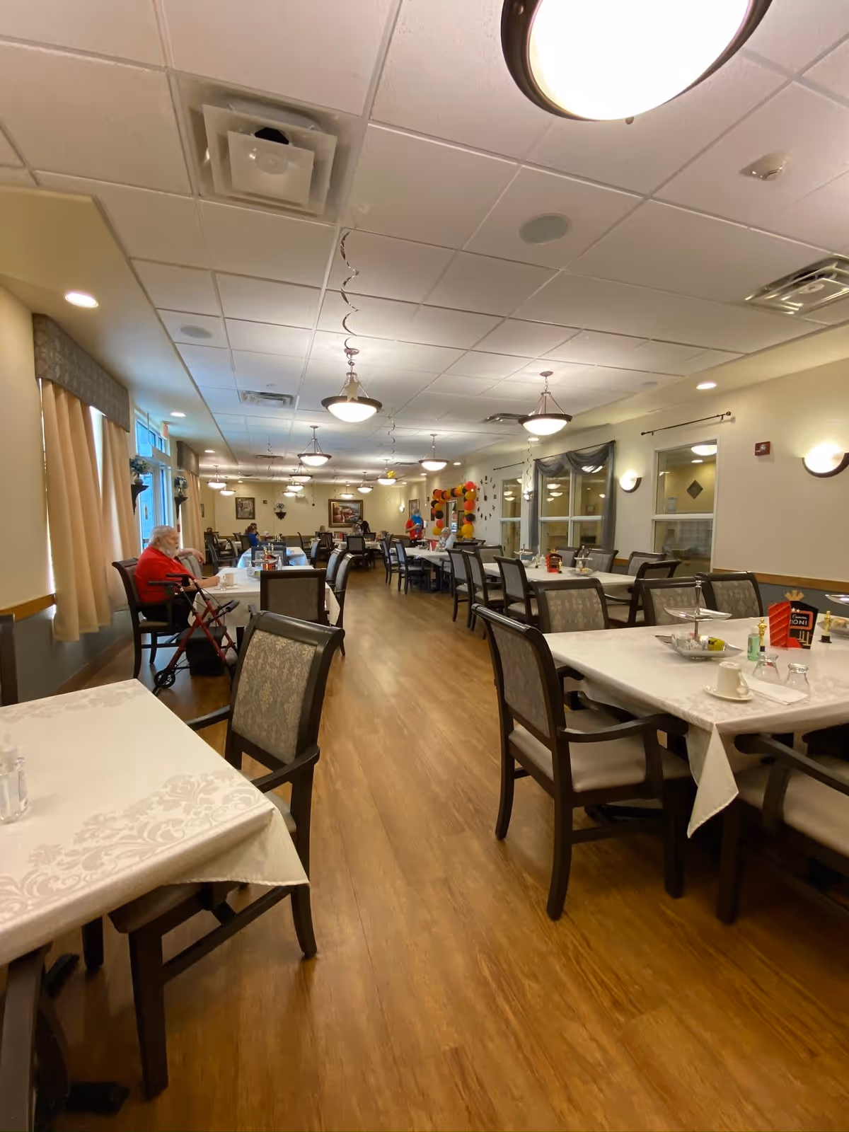 Spacious dining room with rows of tables and chairs, hardwood floor, ceiling lights, and a few residents seated near the windows.