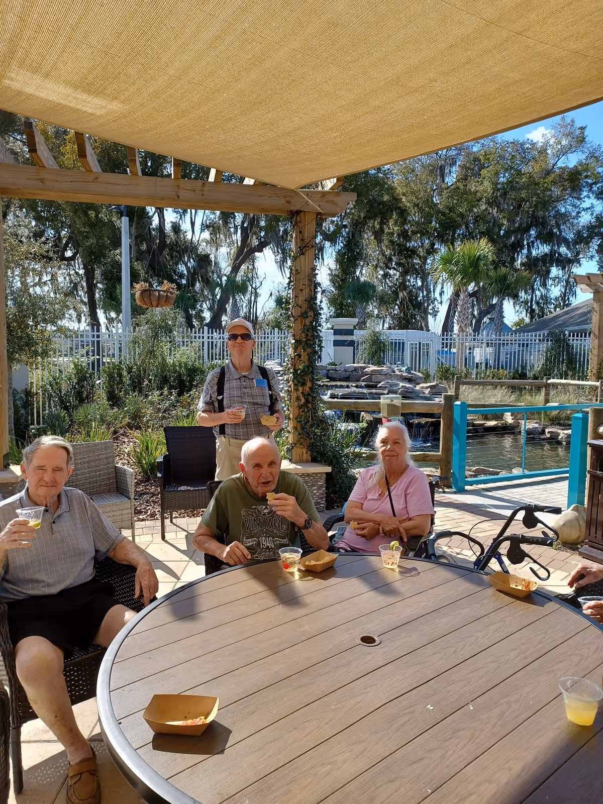 Four elderly people sitting and standing around a round outdoor table under a shade canopy, enjoying drinks and snacks. The setting is a garden area with greenery, a small waterfall feature, and a white fence in the background. One person is in a wheelchair and another has a walker nearby.