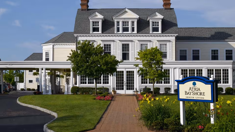 Front exterior view of Atria Bay Shore senior living facility, featuring a large white building with multiple windows, a gray roof, a brick walkway leading to the entrance, green lawn, trees, and a sign with the facility's name surrounded by yellow flowers.