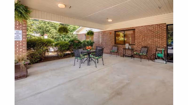 Covered outdoor patio area with a round table and four chairs with green cushions, two additional chairs against a brick wall, potted plants, hanging ferns, and a garden hose reel. There is a 'No Smoking on Front Porch' sign on a brick column.