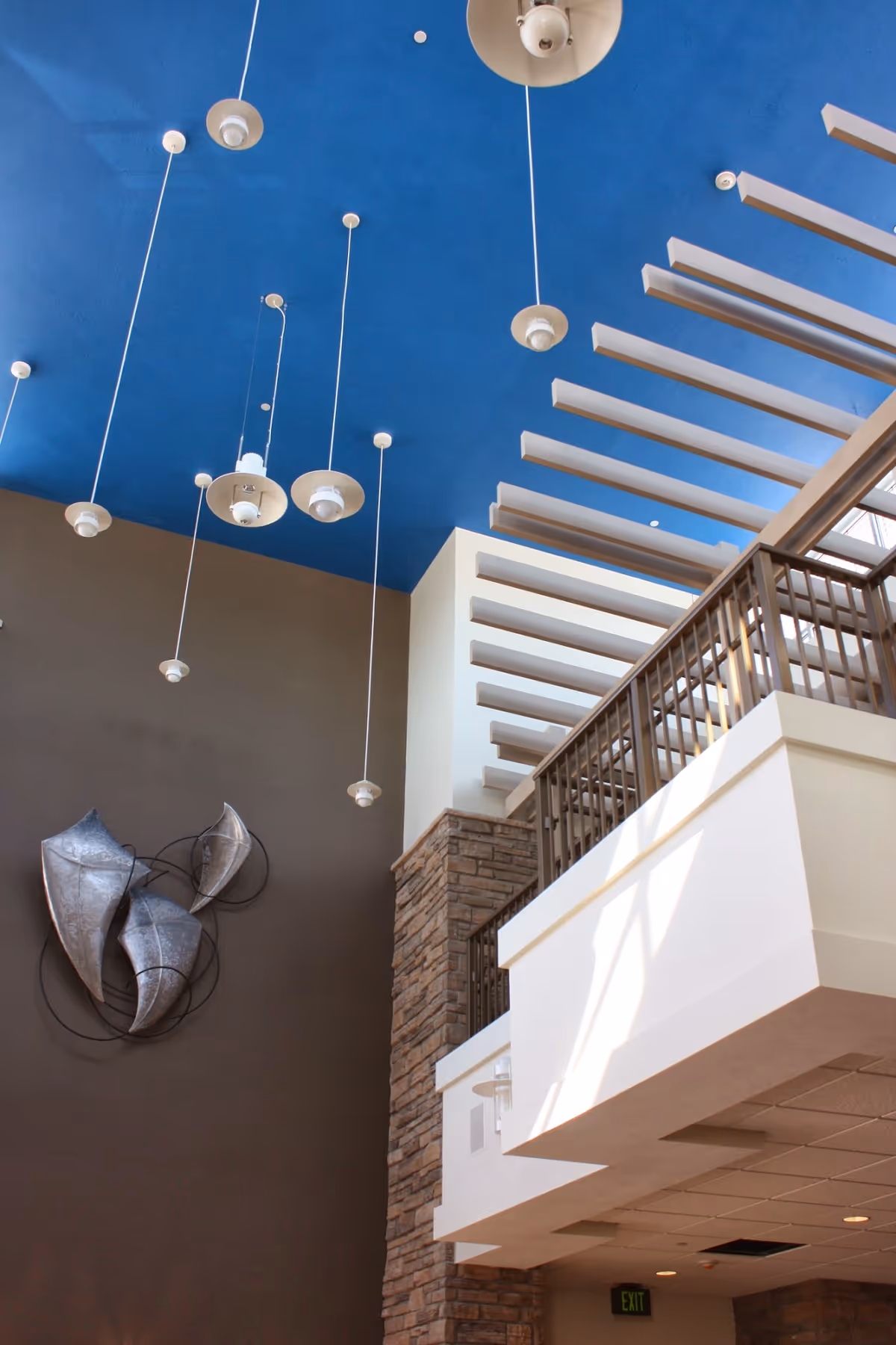 Interior view of a senior living facility with a high ceiling painted blue, multiple hanging pendant lights, a modern metal wall sculpture, stone and white walls, and a balcony with a wooden railing.