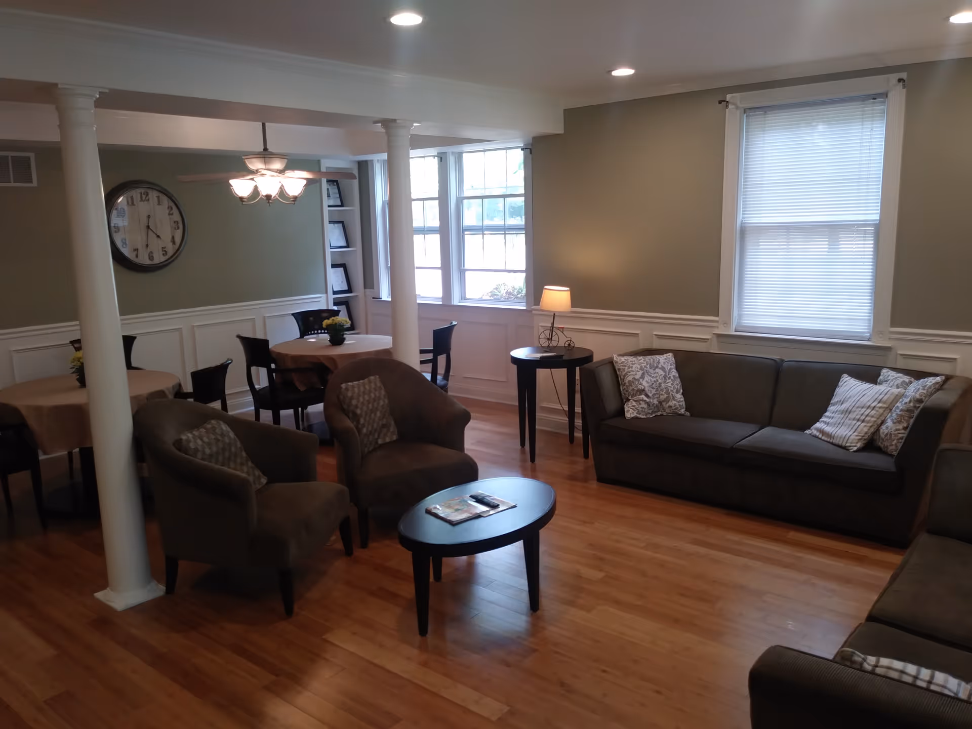 A cozy living room area with wooden flooring, two brown armchairs with patterned cushions, a dark brown sofa with three cushions, a small oval coffee table with magazines and a remote control, and two round tables with chairs in the background. The room has large windows with blinds, a wall clock, a ceiling fan with lights, and a small side table with a lamp.