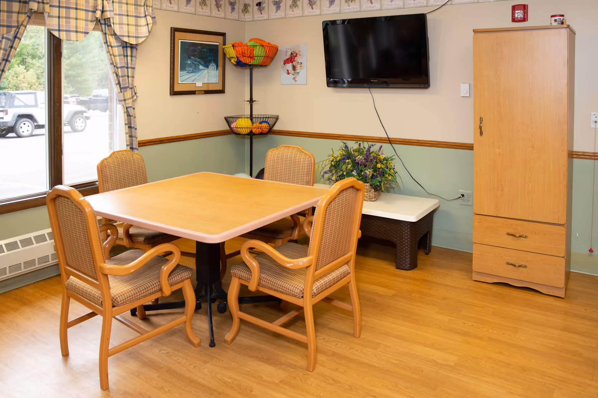 A small dining area with a square wooden table surrounded by four cushioned chairs with wooden frames. The room has light green and beige walls with a wooden chair rail. A flat-screen TV is mounted on the wall above a small table with a flower arrangement. A tall wooden cabinet stands to the right, and a corner fruit basket stand holds colorful artificial fruit. A window with plaid curtains looks out onto a parking lot with vehicles visible outside.
