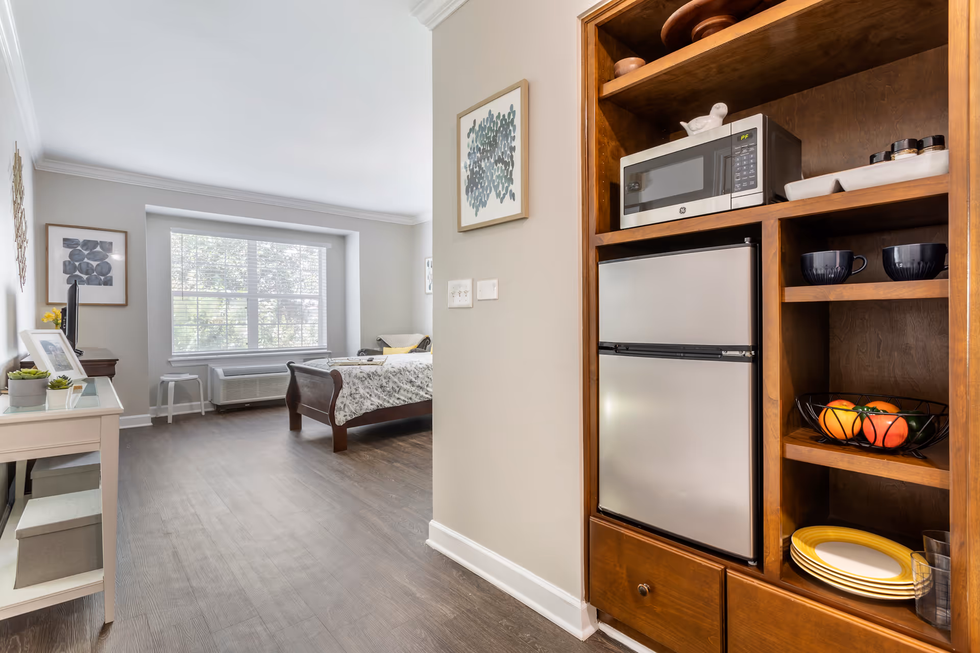 Interior view of a senior living facility room at Brookdale The Heights showing a small kitchenette area with a microwave, mini refrigerator, shelves with cups, plates, and a fruit basket on the right. The room extends into a bedroom area with a bed, a window with blinds, a small table, and framed artwork on the walls.