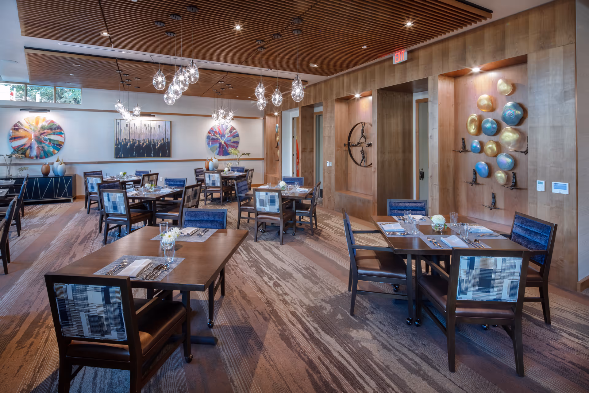 A modern dining room with multiple wooden tables and chairs arranged neatly. The room features a wooden slatted ceiling with hanging pendant lights, colorful abstract wall art, and decorative vases on a sideboard. The floor has a patterned carpet and the walls are adorned with artistic sculptures and paintings.