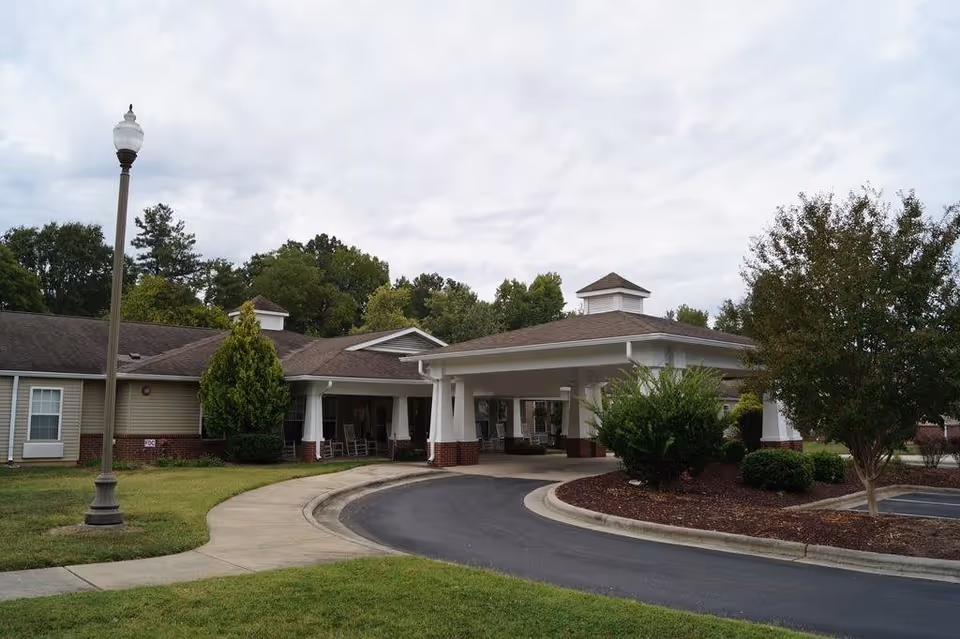 Front entrance and covered porte-cochère of a single-story senior living facility with a curved driveway, landscaping, and a lamppost.