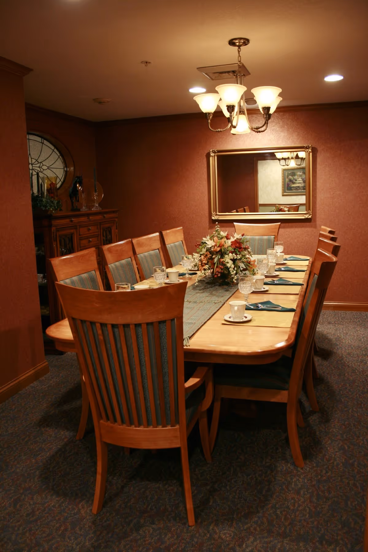 A formal dining room with a long wooden table set for eight people. The table has a floral centerpiece, placemats, cups, glasses, and napkins. The room features a chandelier overhead, a large mirror on the wall, and a wooden cabinet with decorative items in the corner.