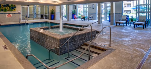 Indoor swimming pool area with a central raised fountain feature, steps leading into the pool, and seating areas with chairs and tables near large windows letting in natural light.