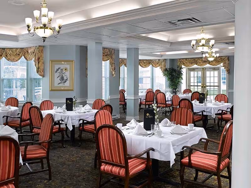 A formal dining room with multiple tables covered in white tablecloths, each set with white napkins, cups, and menus. The chairs have red and orange striped upholstery. The room has large windows with decorative valances, chandeliers hanging from the ceiling, and framed artwork on the walls.