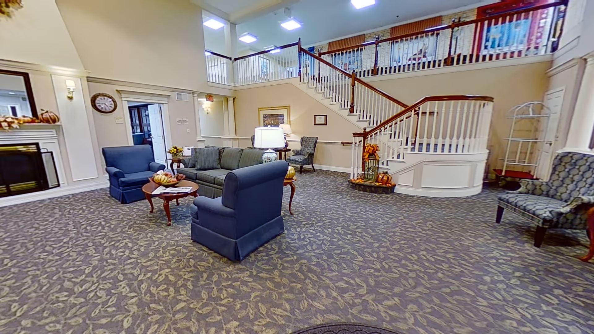 A spacious living room area with patterned carpet, a green sofa, two blue armchairs, a wooden coffee table with decorative pumpkins, and a fireplace on the left. There is a staircase with white railings leading to an upper floor, and additional seating including a patterned chair on the right. The room is well-lit with ceiling lights and wall sconces, and decorated with autumn-themed items.