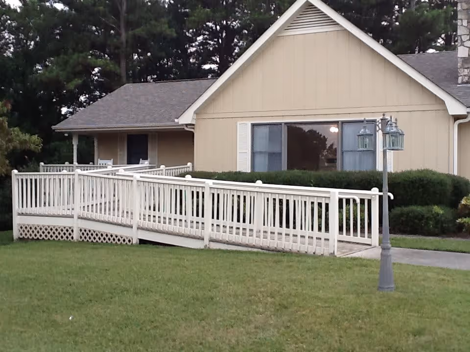 Exterior view of a beige single-story building with a white wheelchair ramp leading to the entrance. The building has a large window with white shutters and is surrounded by green bushes and a well-maintained lawn. There is a decorative street lamp with two lanterns near the ramp.