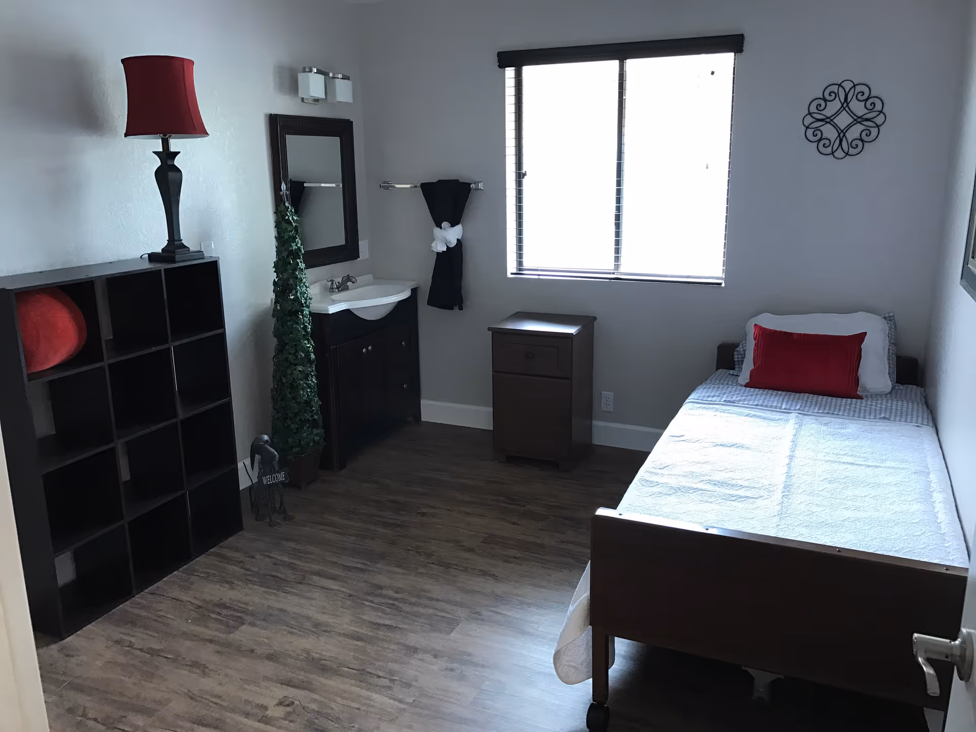 A simple bedroom with a single bed covered in white bedding and a red pillow, a wooden nightstand, a window with blinds, a small sink with a mirror above it, a black shelving unit with a red lamp on top, and a decorative green plant next to the sink.