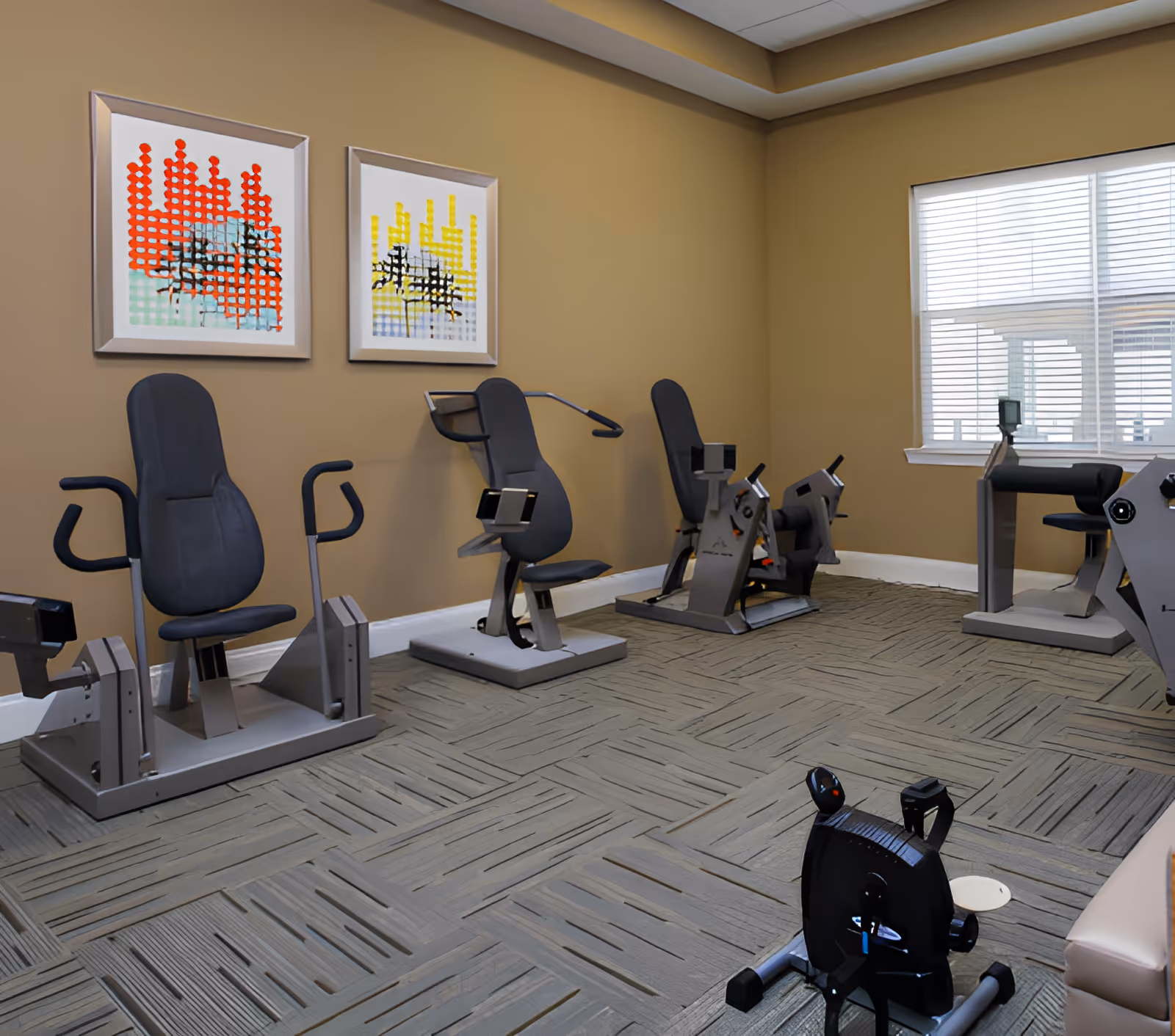 Exercise room with multiple seated cardio machines, framed artwork on a beige wall, and a window letting in light.