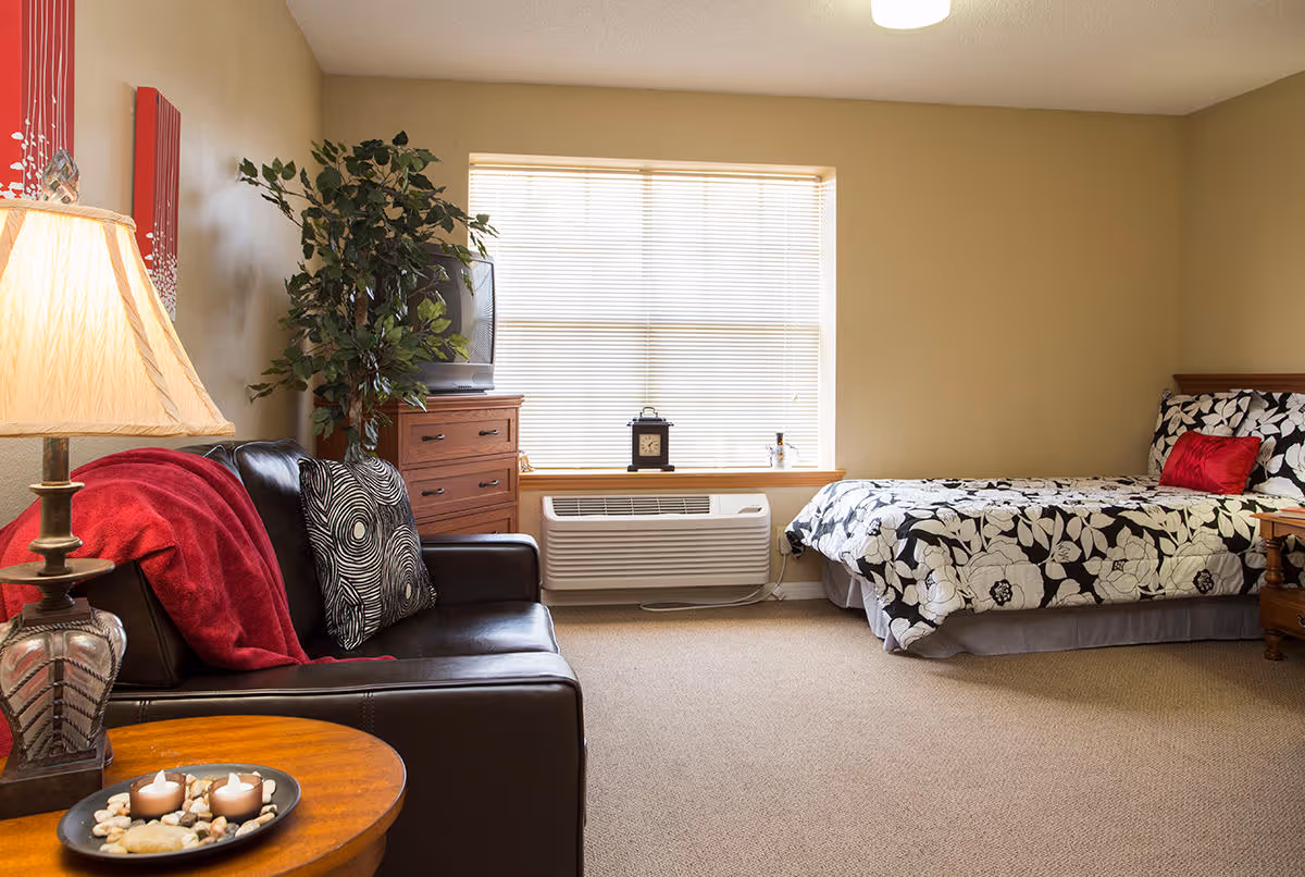 A cozy bedroom in Bradfield Terrace featuring a bed with black and white floral bedding and a red pillow, a dark leather couch with patterned cushions and a red throw, a wooden dresser with a TV on top, a large window with blinds, a potted plant, and a side table with a lamp and decorative candles.