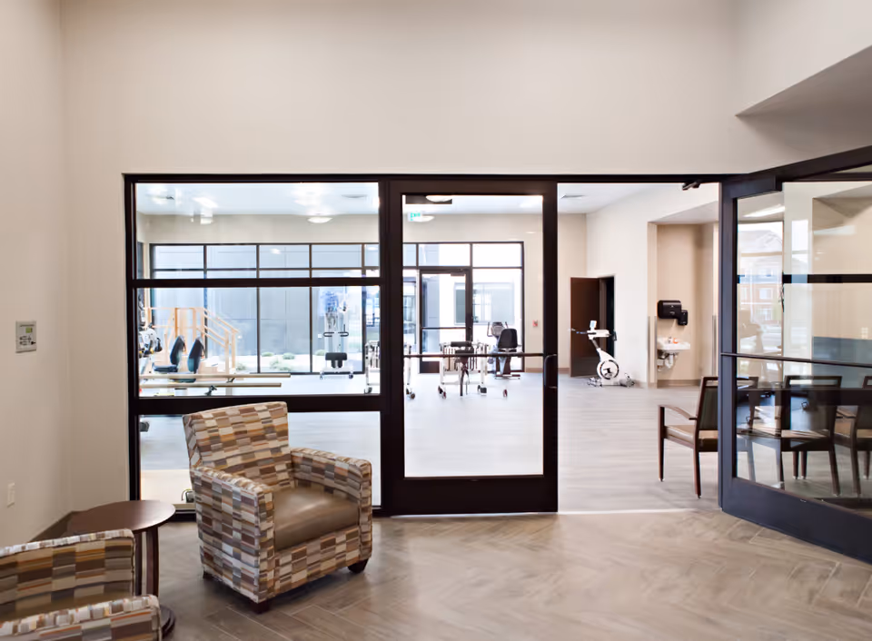 View of a modern interior space with two patterned armchairs and a small round table in the foreground, separated by glass doors from a bright room with exercise equipment and a stationary bike in the background.