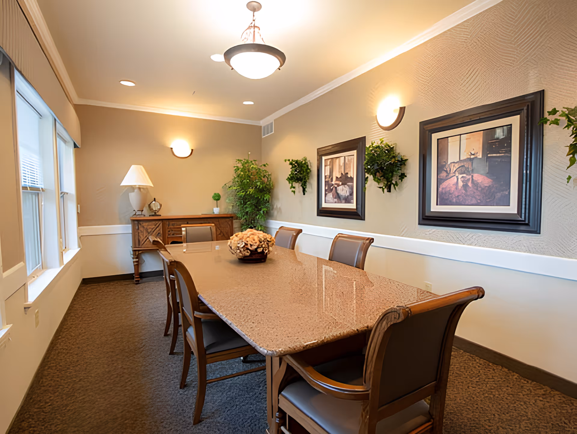A well-lit dining room with a long rectangular table surrounded by six wooden chairs with cushioned seats. The table has a decorative centerpiece with flowers. The room features beige walls with white trim, two framed pictures on the wall, wall-mounted light fixtures, a sideboard with a lamp and small plants, and a large window letting in natural light.