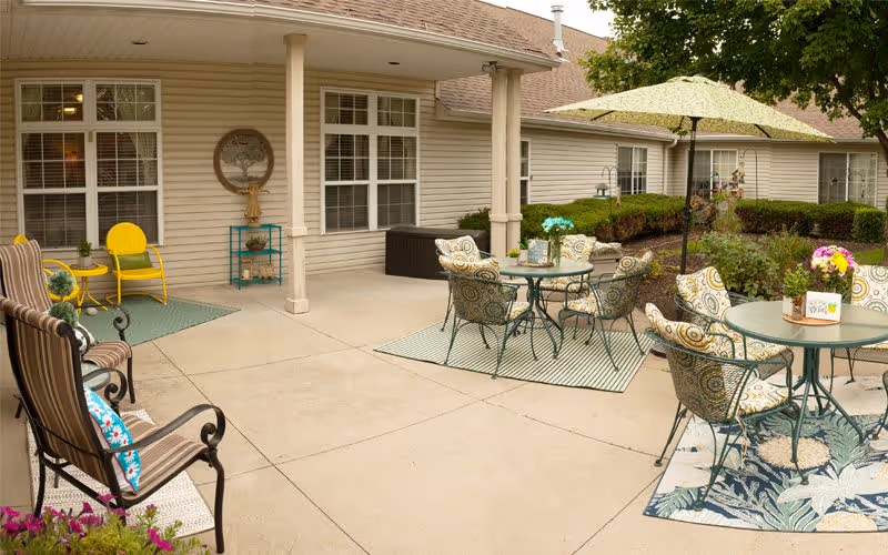 Sunny outdoor patio and courtyard with multiple tables, cushioned chairs, umbrellas, potted plants, and the building exterior.