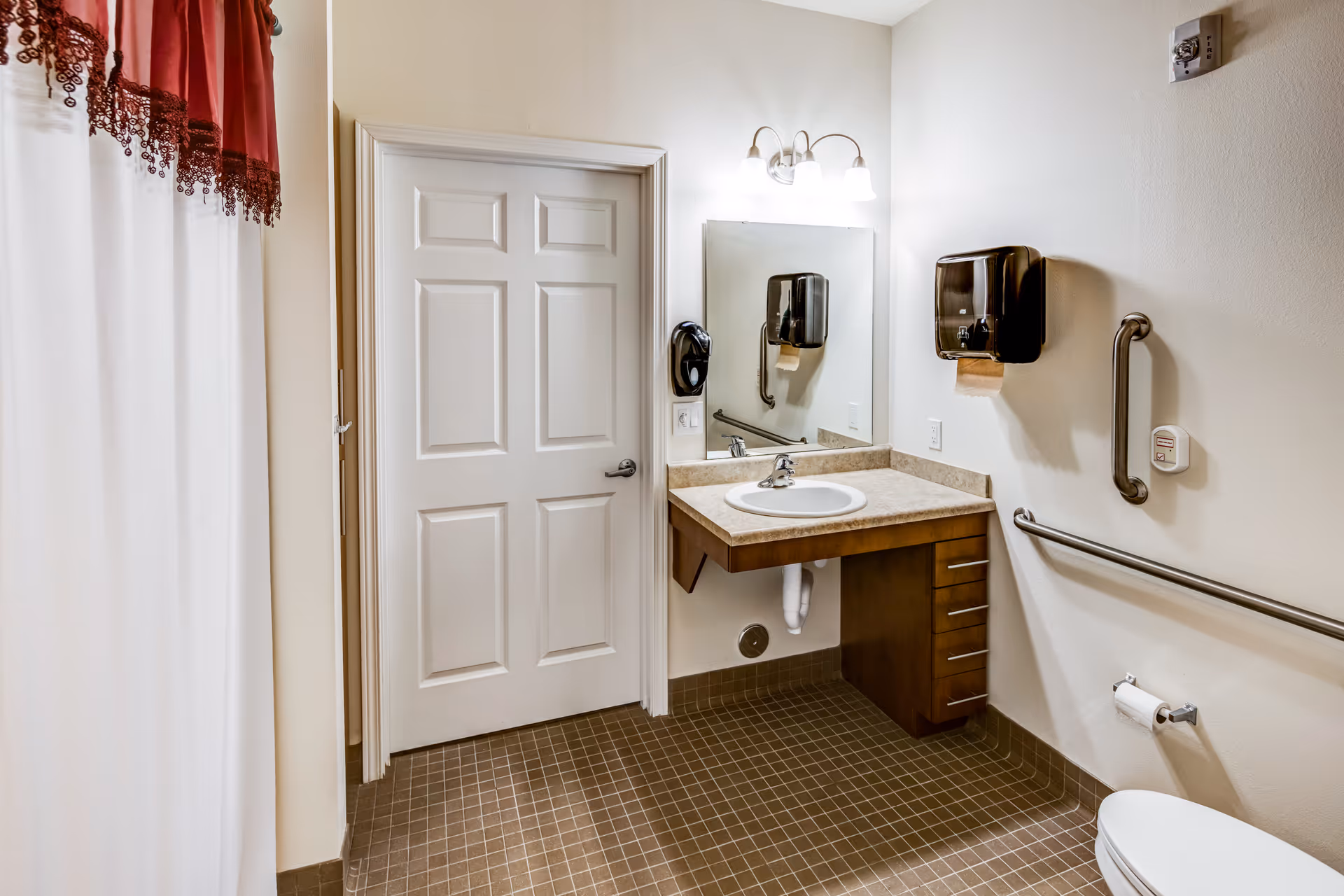 A clean and accessible bathroom featuring a white door, a wall-mounted sink with a countertop and drawers underneath, a large mirror above the sink, a soap dispenser, grab bars on the walls, a toilet with a toilet paper holder, and a shower curtain with a red valance.